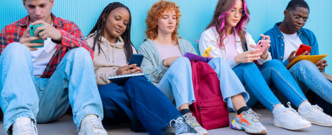 A diverse group of five young students sit together on the floor, smiling and interacting on their smartphones against a blue background.