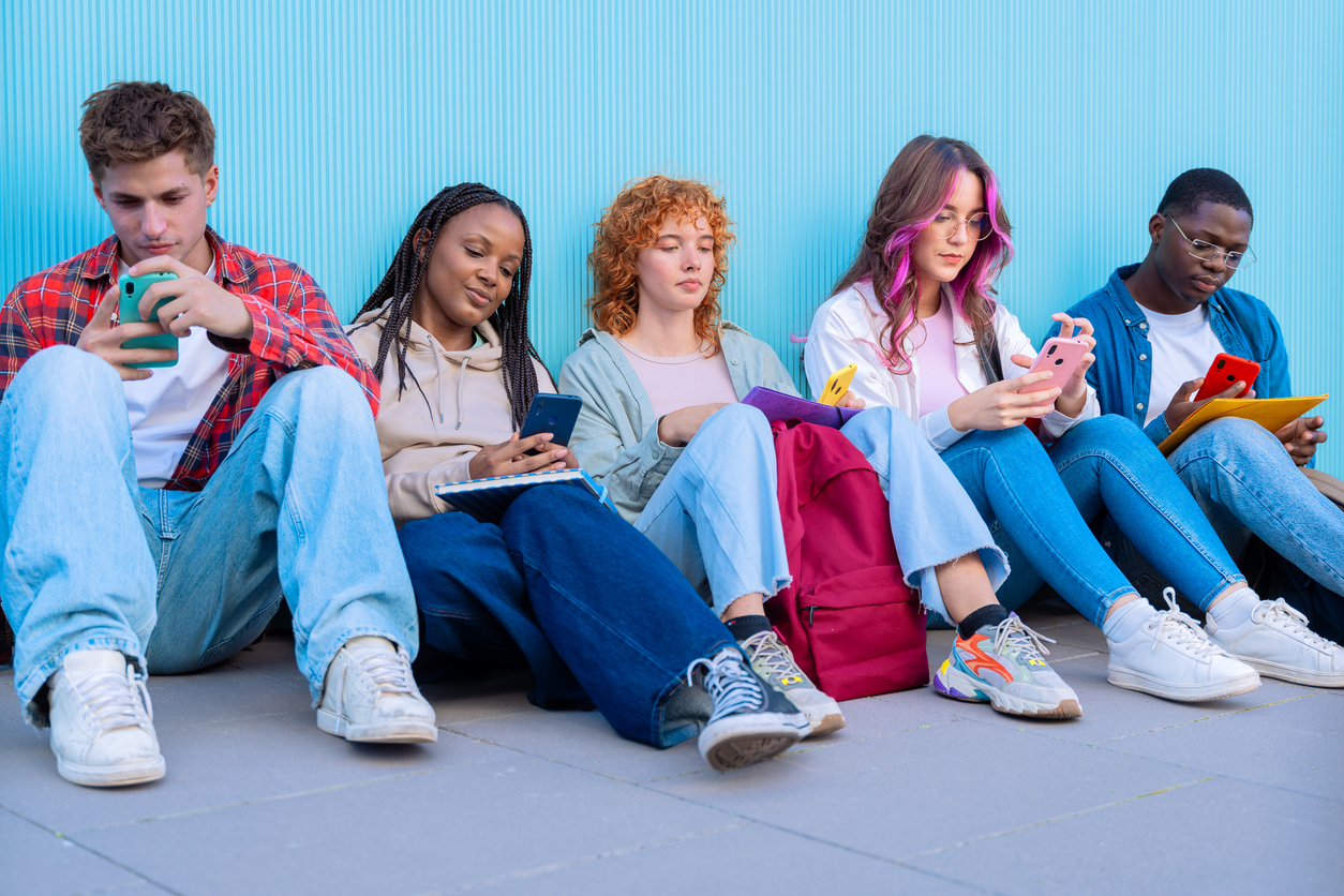 A diverse group of five young students sit together on the floor, smiling and interacting on their smartphones against a blue background.